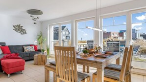 Un coin repas avec une table et des chaises en bois, un pouf rouge, un canapé gris avec des coussins rouges et une grande fenêtre donnant sur des immeubles et une plage.