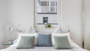 Une chambre avec un grand lit, deux tables de chevet, une fenêtre donnant sur un bâtiment blanc et une petite plante en pot posée sur le rebord de la fenêtre.