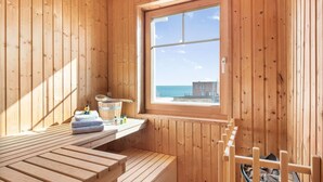 Un sauna en bois, avec un banc, une fenêtre donnant sur la mer et une étagère en bois où sont posées des serviettes et des bouteilles.