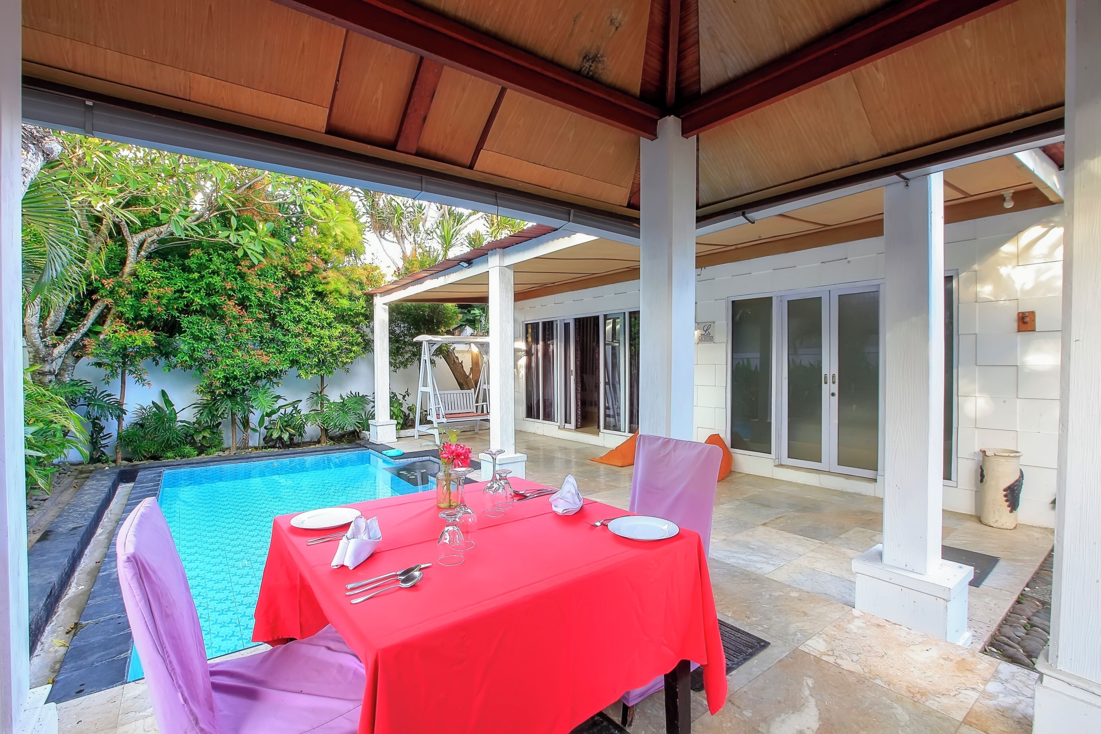 A covered outdoor dining area with a pool, white columns, and lush greenery.