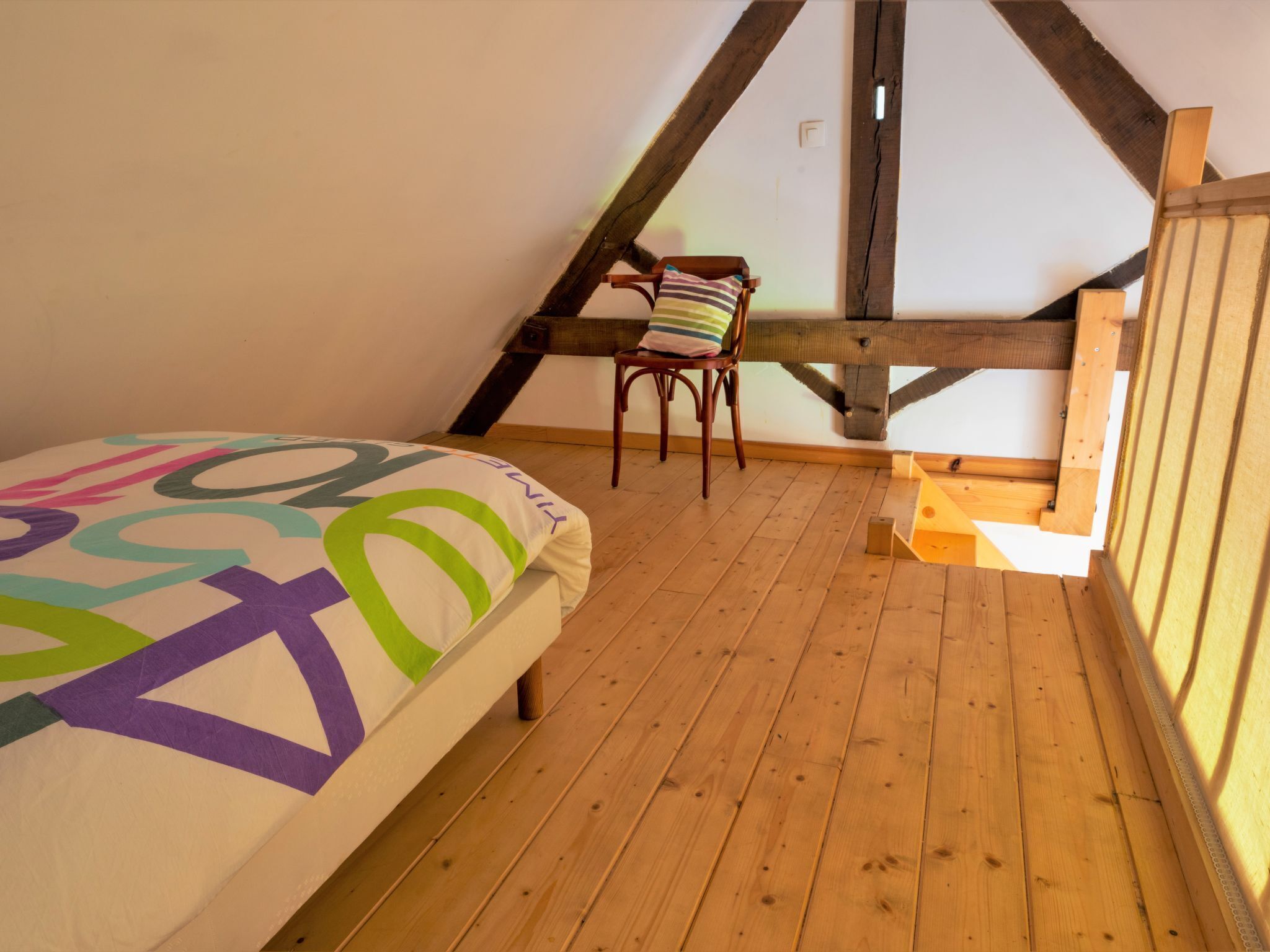 A bedroom with a wooden floor, a bed with a colorful bedspread, a chair, and a slanted ceiling with wooden beams.