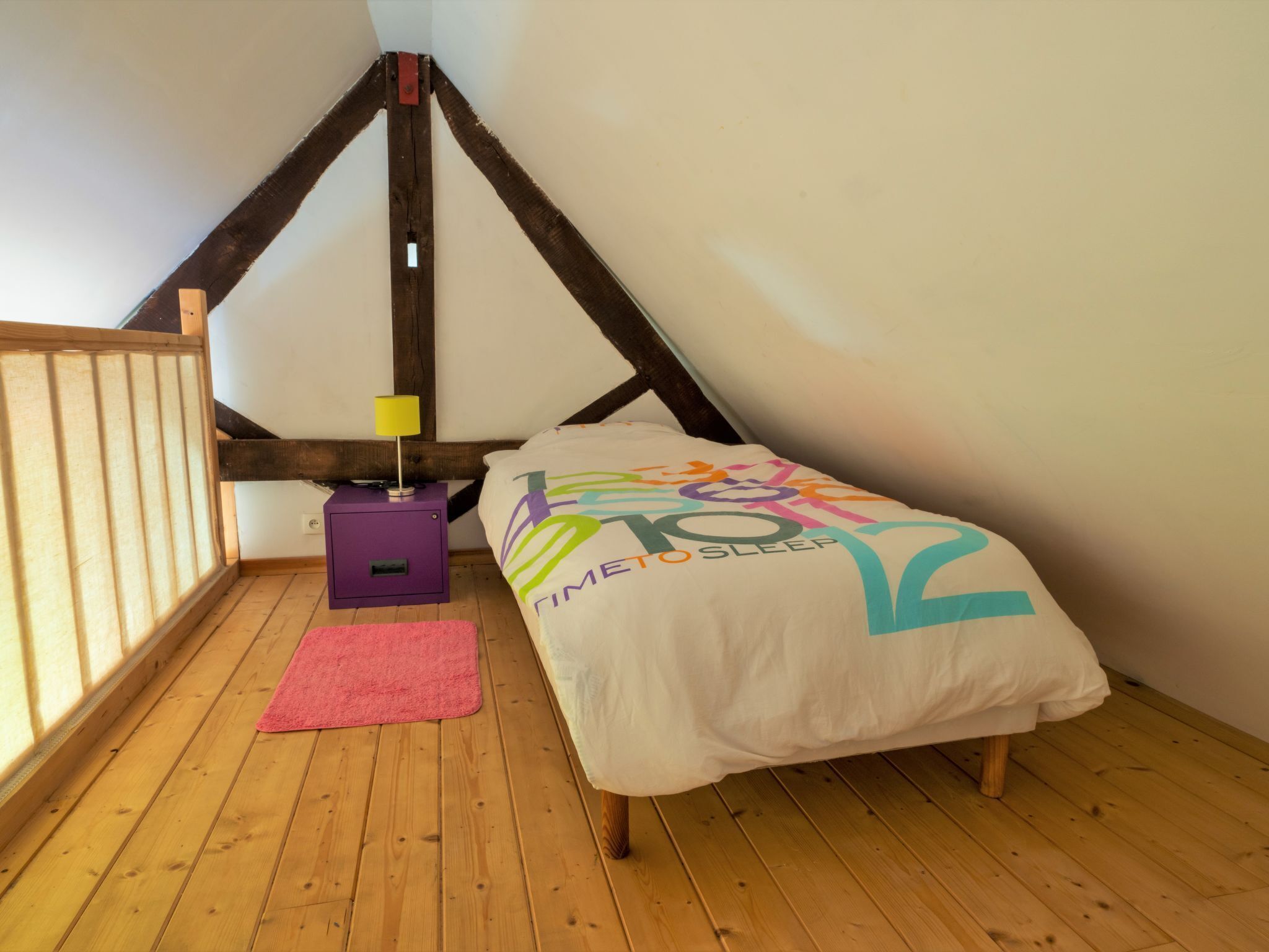 Une chambre avec un plafond en pente, un lit recouvert d’une housse colorée sur laquelle est écrit « Il est l’heure de dormir », une table de chevet violette avec une lampe, et un tapis rose.
