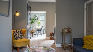 A living room with a yellow armchair, a blue sofa, a coffee table with board games, and a dining area in the background.