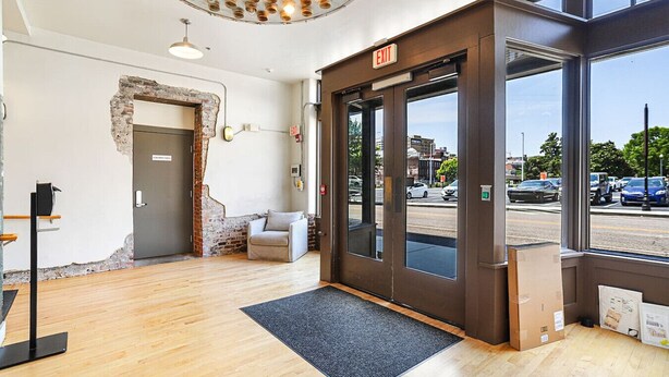 Een modern interieur met een houten vloer, een grijze bank, een zwarte deur met een 'EXIT'-bord en uitzicht op de straat door grote ramen.