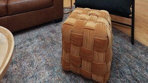 Een moderne woonkamer met een bruine leren bank, een zwarte fauteuil en een geweven pouf. Er staat een vloerlamp, een eettafel met stoelen en een zichtbare keuken.