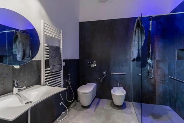 Une salle de bain moderne avec un grand miroir, un lavabo blanc, un bidet, des toilettes et une douche séparée par une paroi en verre.