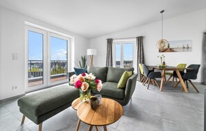 Una sala de estar moderna con un sofá gris, una mesa de centro redonda de madera y un jarrón con flores. Hay un comedor con una mesa y sillas, y un balcón con vistas a casas.