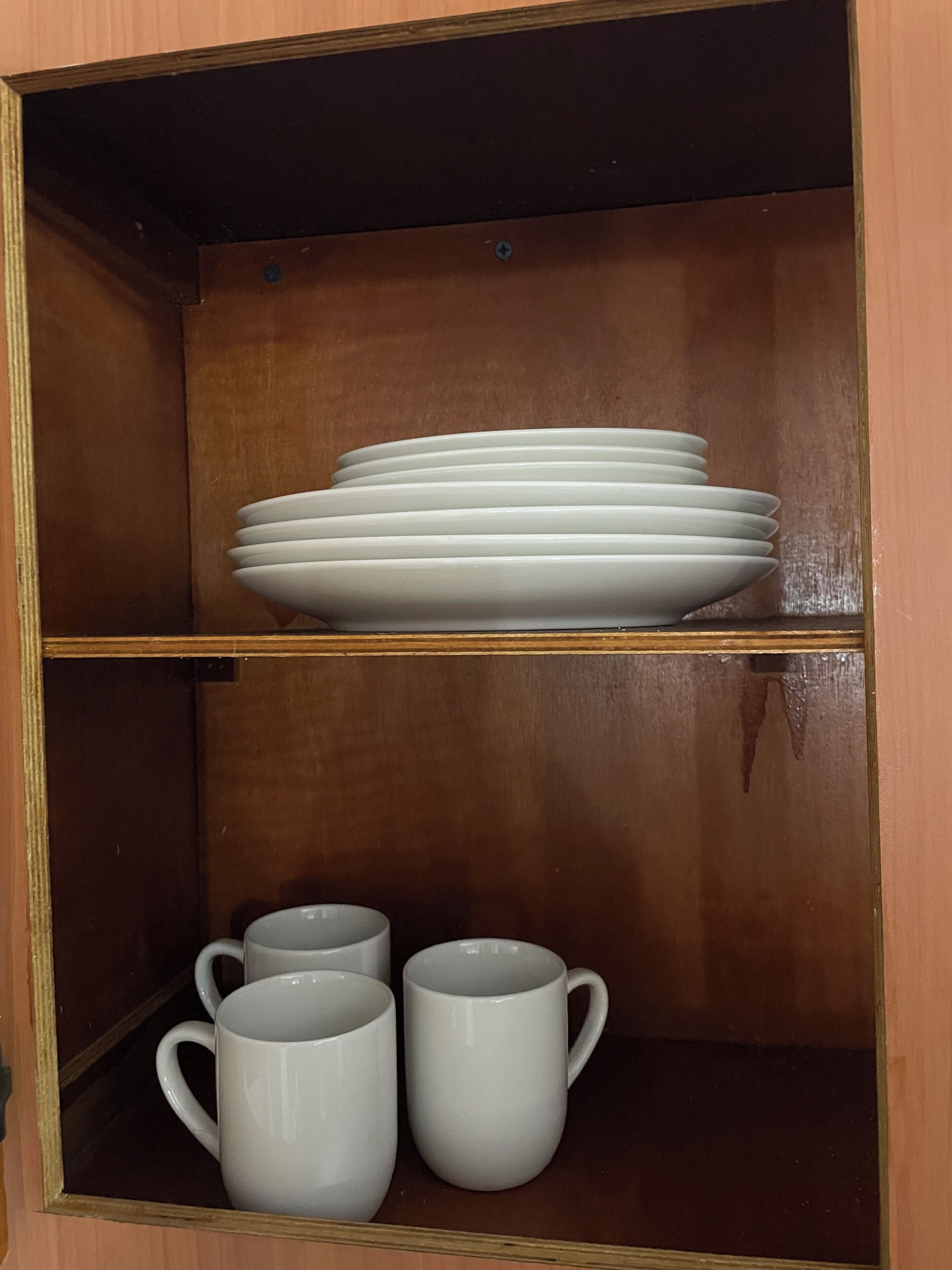 Un meuble en bois à deux étagères ; l’étagère supérieure contient une pile d’assiettes blanches, et l’étagère inférieure abrite trois tasses blanches.