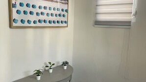 Une pièce décorée d’un tableau représentant une scène de plage, une petite table sur laquelle sont posées deux plantes en pot, et une fenêtre avec des stores.