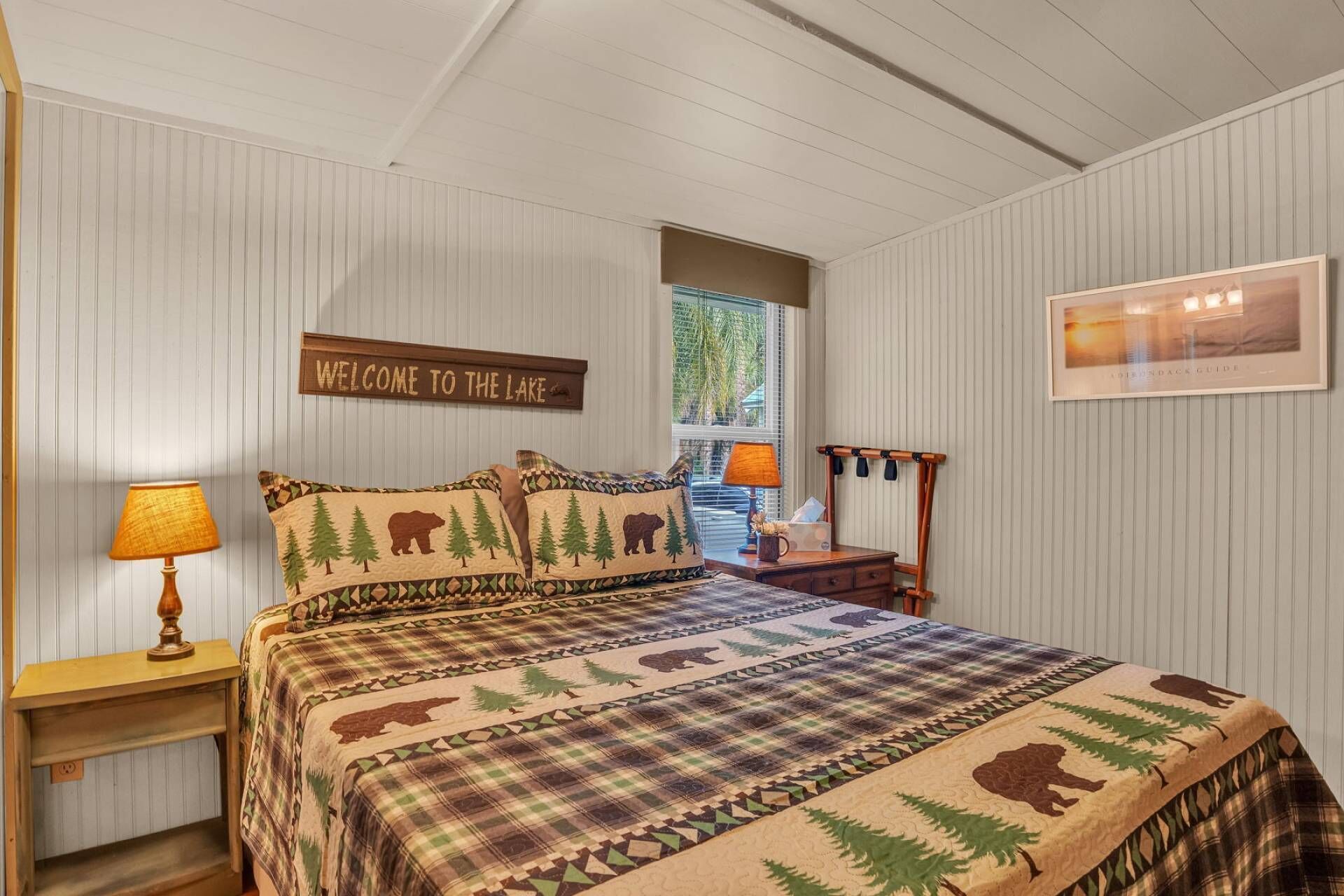 A bedroom with a plaid bedspread featuring bear and tree patterns, a 'Welcome to the Lake' sign, and a framed picture on the wall.