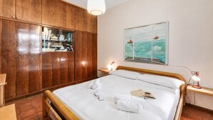 A hotel room with a wooden wardrobe, a bed with white linens, a bedside table with a lamp, and a painting of a ship on the wall. - Appartamento Alessandra: Terrace and Lake View, Laveno Mombello, Italy (Laveno Mombello)