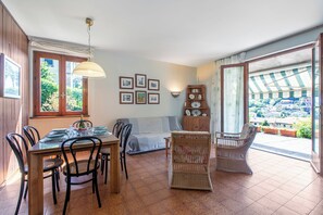 A dining area with a table set for four, a sofa, a wicker chair, and a view of the outside through a large window. - Appartamento Alessandra, Laveno Mombello, Italy (Laveno Mombello)