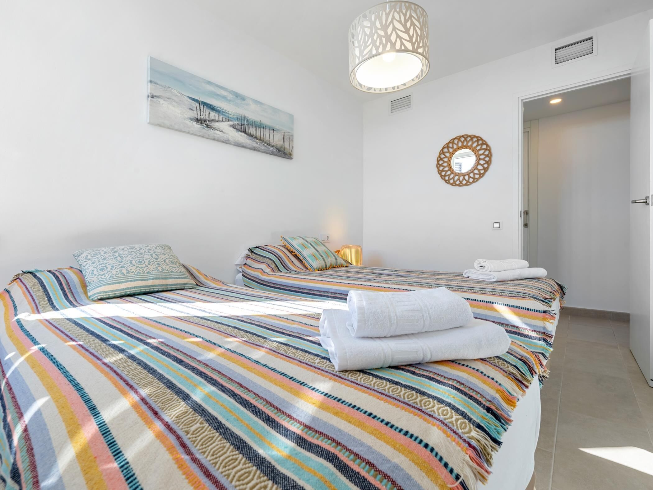 Habitación de hotel con dos camas, ropa de cama a rayas, un cuadro en la pared, un espejo decorativo y una puerta que da a otra habitación.