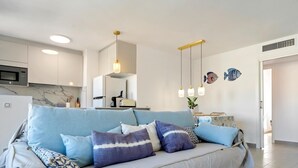 A modern living room with a light blue sofa, patterned cushions, and a kitchen in the background. - Apartment 250m beach with pool and parking (Torredembarra)