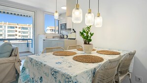A dining area with a table set for four, surrounded by wicker chairs, a potted plant, and a view of the cityscape through large windows. - Apartment 250m beach with pool and parking (Torredembarra)