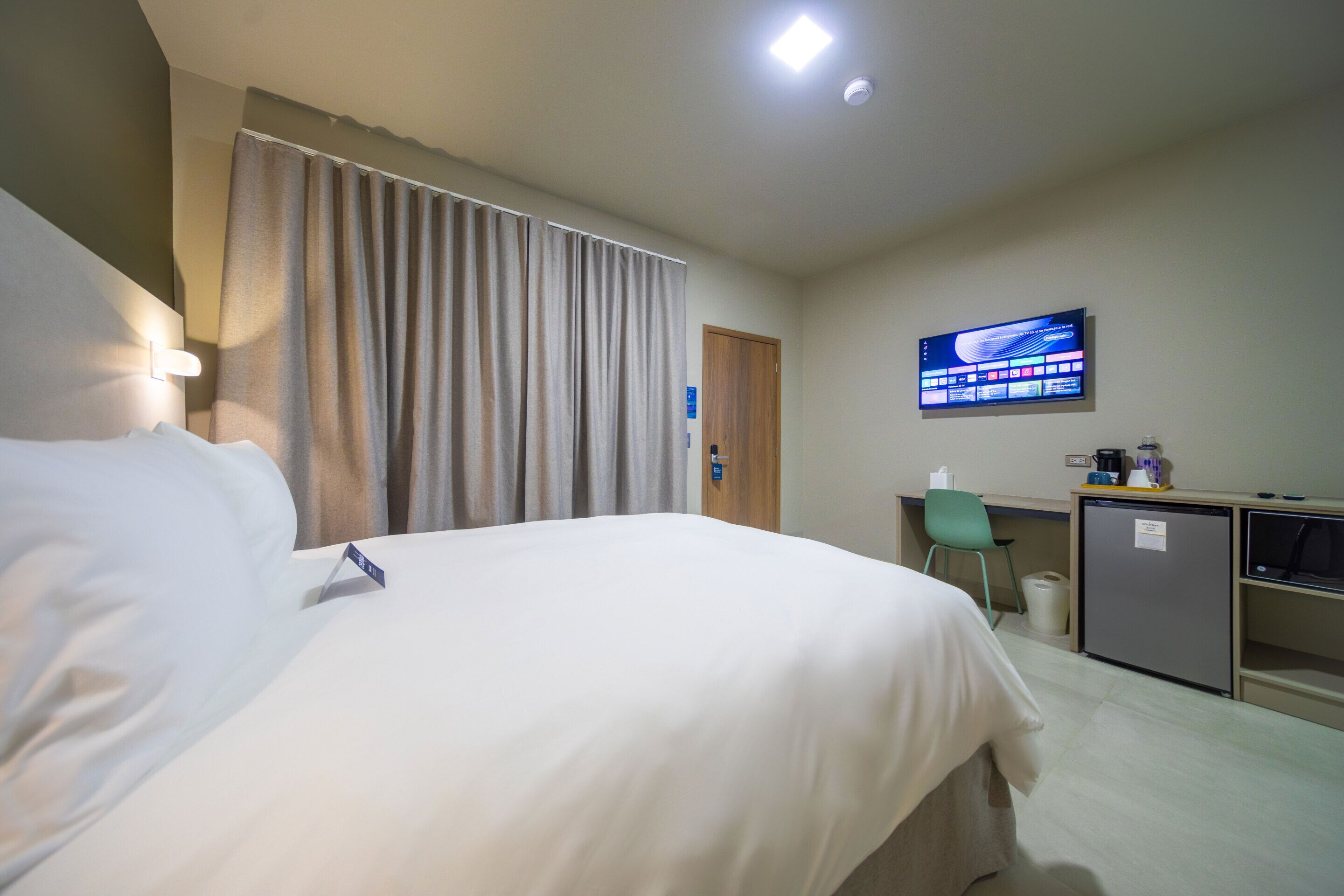 Une chambre d’hôtel avec un grand lit, un bureau avec un ordinateur, une télévision et un petit réfrigérateur.