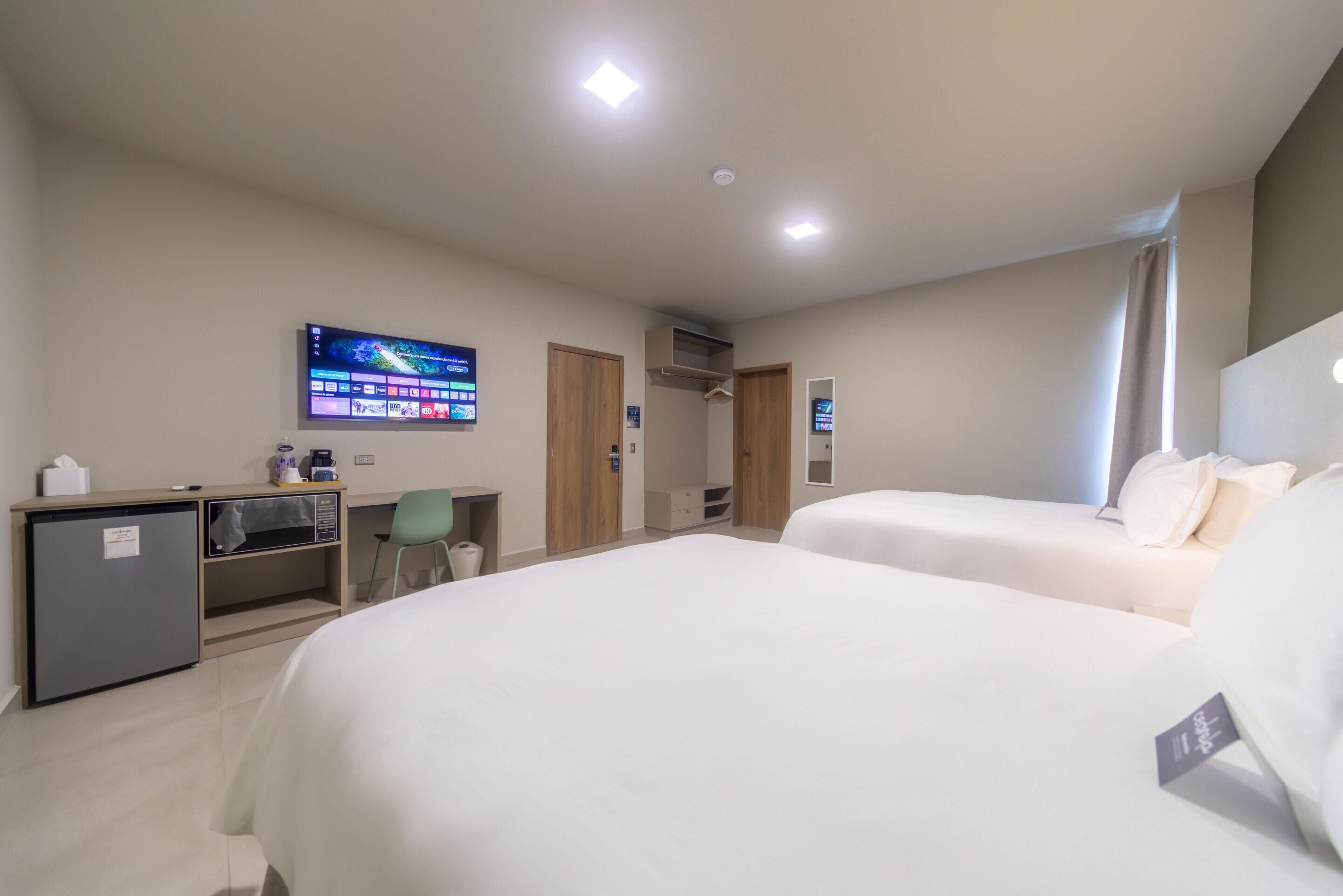 A hotel room with two beds, a desk with a computer, a small fridge, and a TV mounted on the wall.