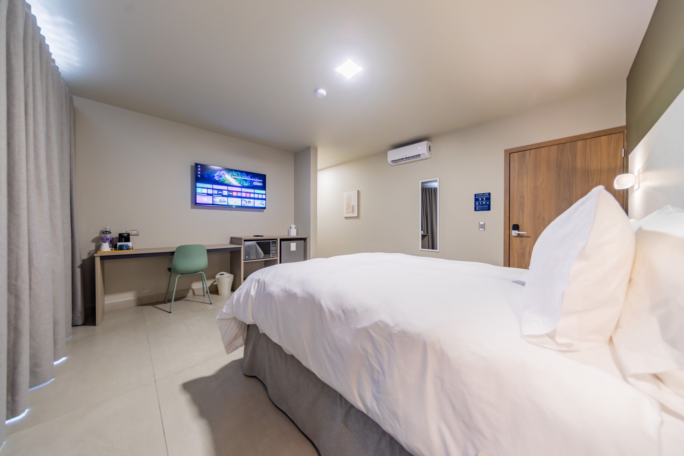 A hotel room with a bed, a desk, a chair, a TV, an air conditioning unit, and a door.