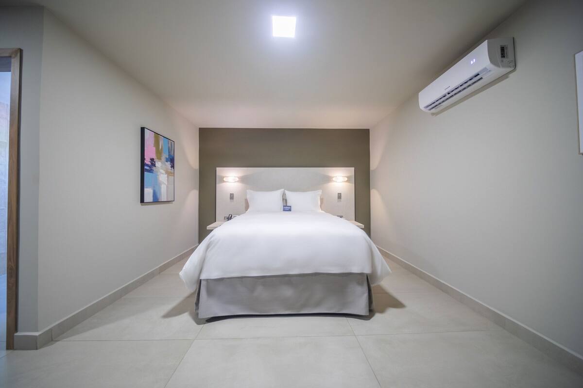 A hotel room with a bed, a headboard with lights, an air conditioner, and a framed picture on the wall.