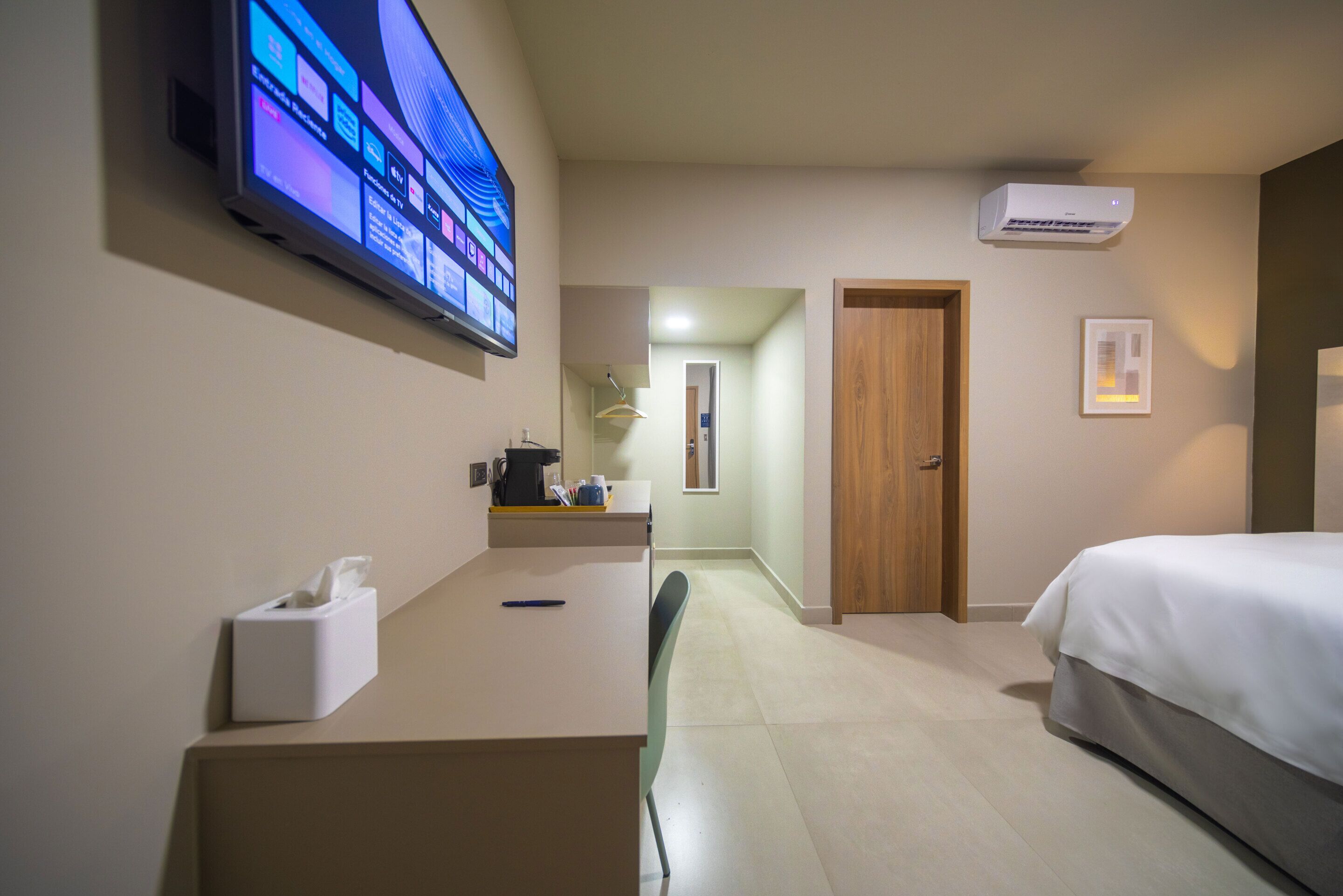 Quarto de hotel com cama, escrivaninha com monitor de computador, cafeteira e televisão fixada na parede.