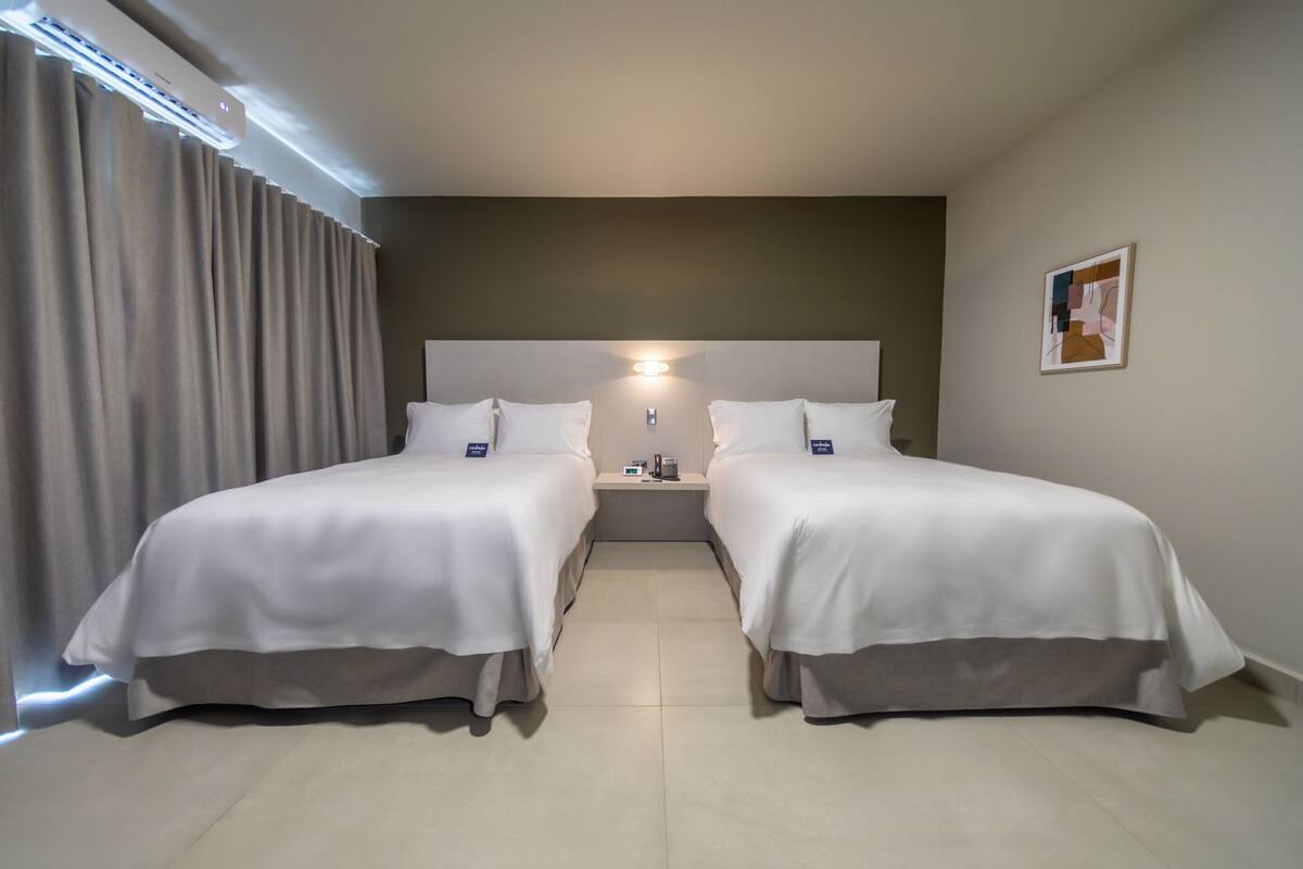 A hotel room with two beds, a nightstand, a wall-mounted light, and a window with curtains.