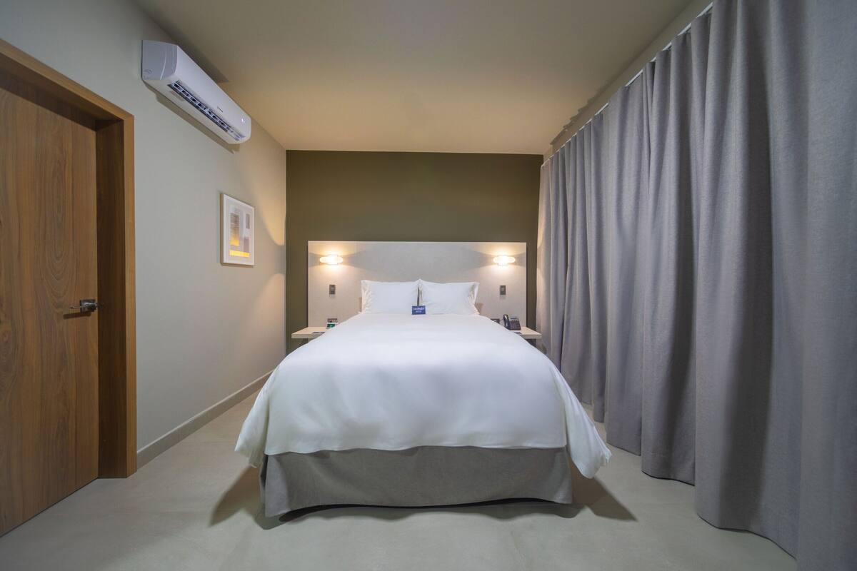 A hotel room with a large bed, an air conditioning unit, a wooden door, and a curtain.