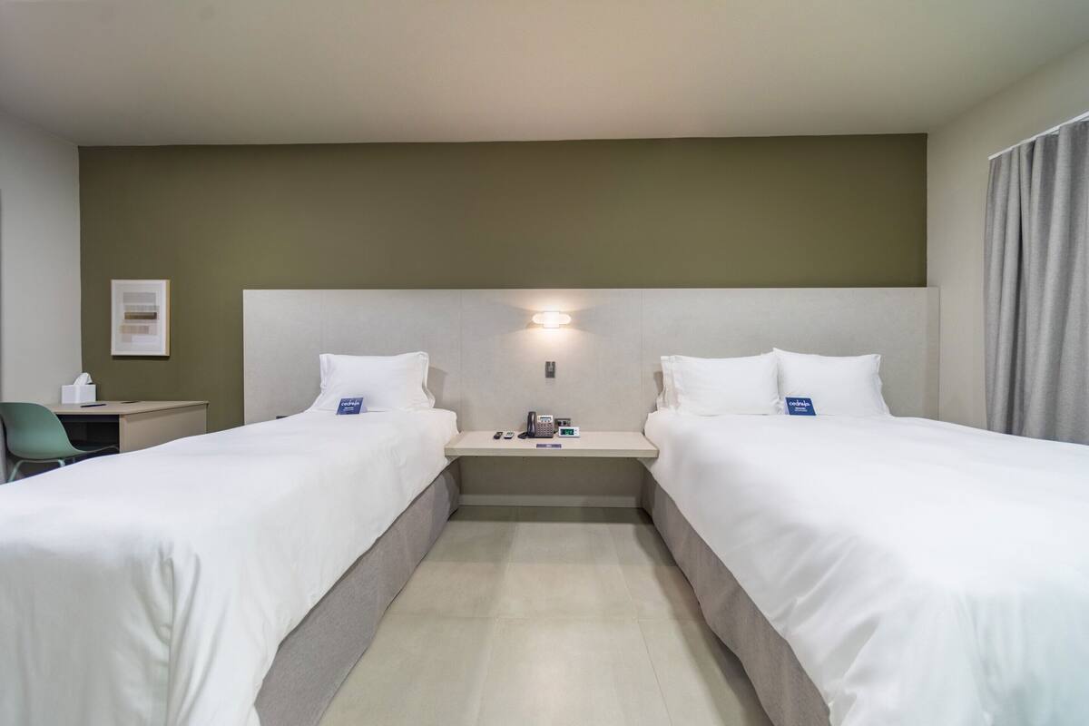 A hotel room with two single beds, a bedside table with a lamp, a chair, and a small desk with a telephone and a tissue box.