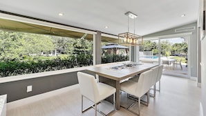 Un comedor moderno con una gran mesa y sillas, una lámpara colgante y vistas a una piscina y follaje exterior.