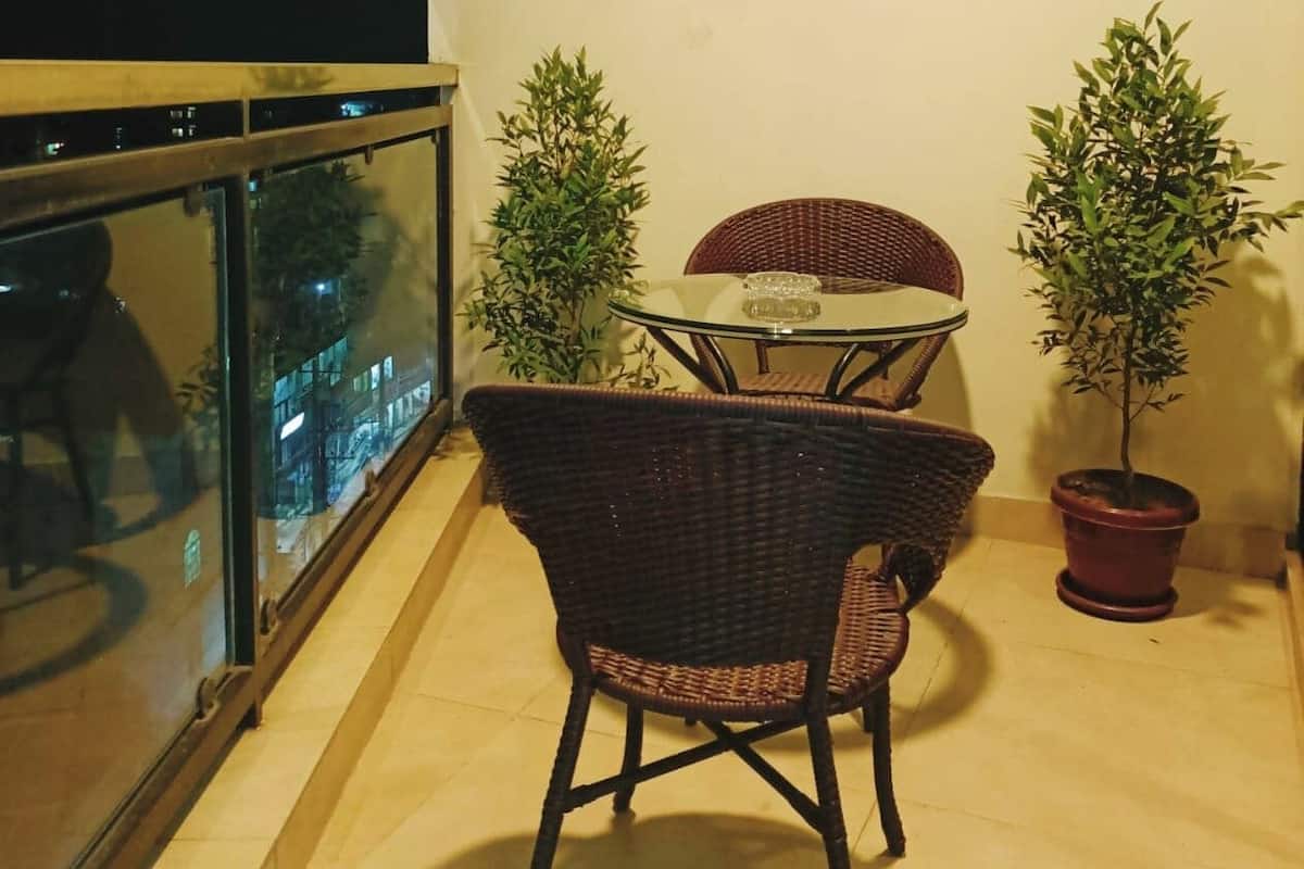 Un balcón con barandilla de vidrio, una silla de mimbre, una mesita redonda y dos macetas con plantas.