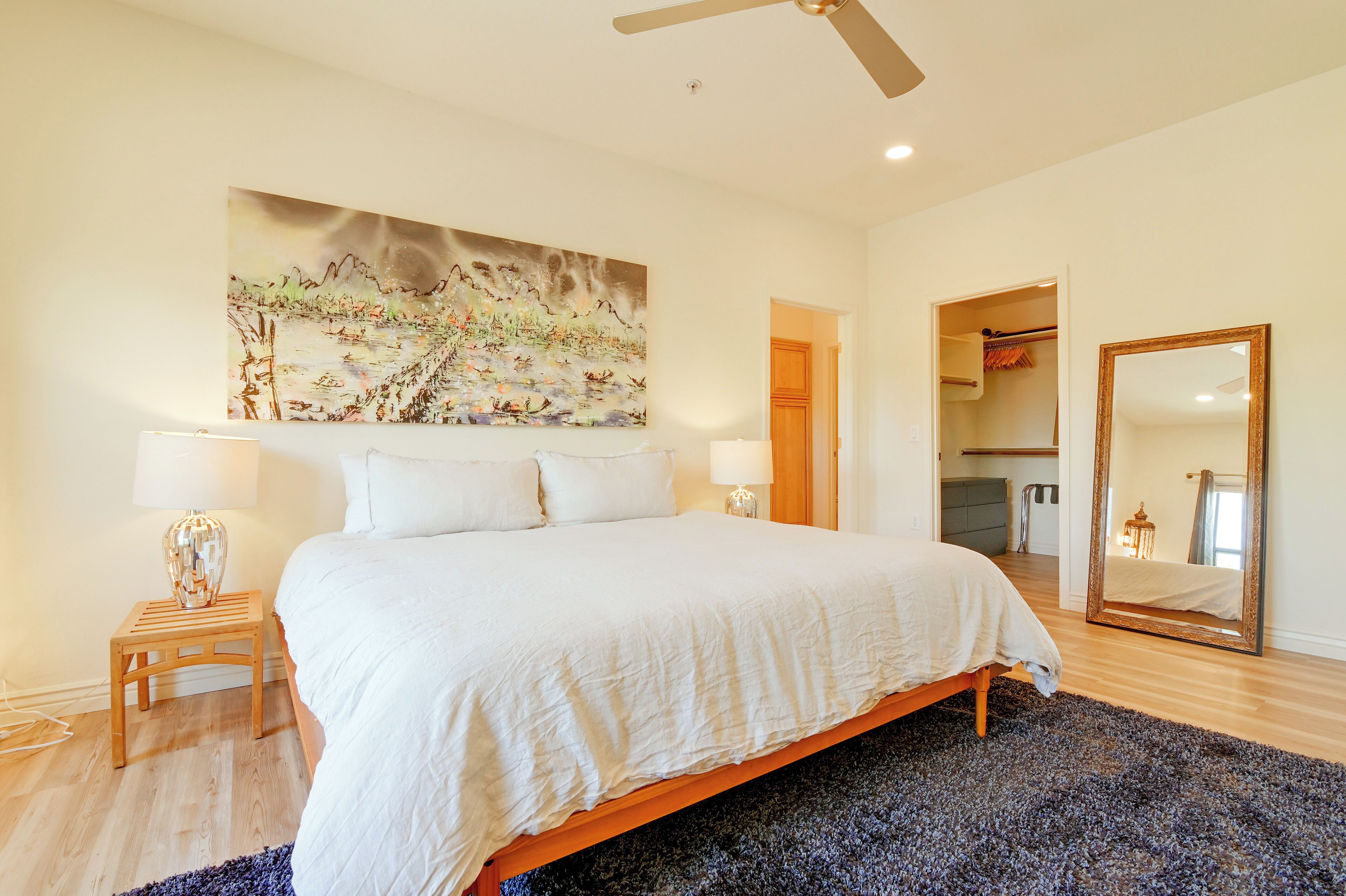 A bedroom with a large bed, two bedside lamps, a ceiling fan, and a large painting on the wall.