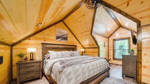 Ein Schlafzimmer mit Holzboden, einem Bett mit gemustertem Überwurf, einem Nachttisch mit Lampe und einem Fenster mit Blick auf Bäume.