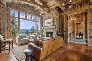 A rustic interior with a stone fireplace, wooden beams, and a large window offering a mountain view. - Luxury 5 bed, 4 bath house I Private I Tranquility (Whitefish)
