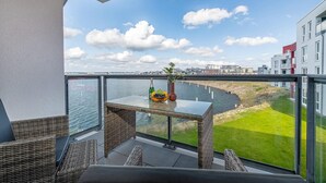 Een balkon met rieten meubilair, een glazen tafel met een fles en fruitschaal, en uitzicht op een rivier en gebouwen.