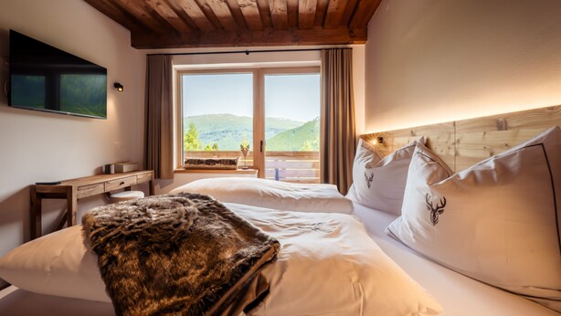 A bedroom with a wooden ceiling, a large window with a view of hills, a bed with white linens, and a television on the wall. - Idyllic holiday apartments for travel groups (Kremsbrücke)