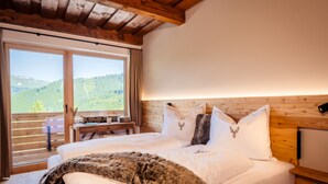 A bedroom with a wooden ceiling, a large window with a view of green hills, a bed with white bedding and a fur throw, and a bedside table with a lamp. - Idyllic holiday apartments for travel groups (Kremsbrücke)