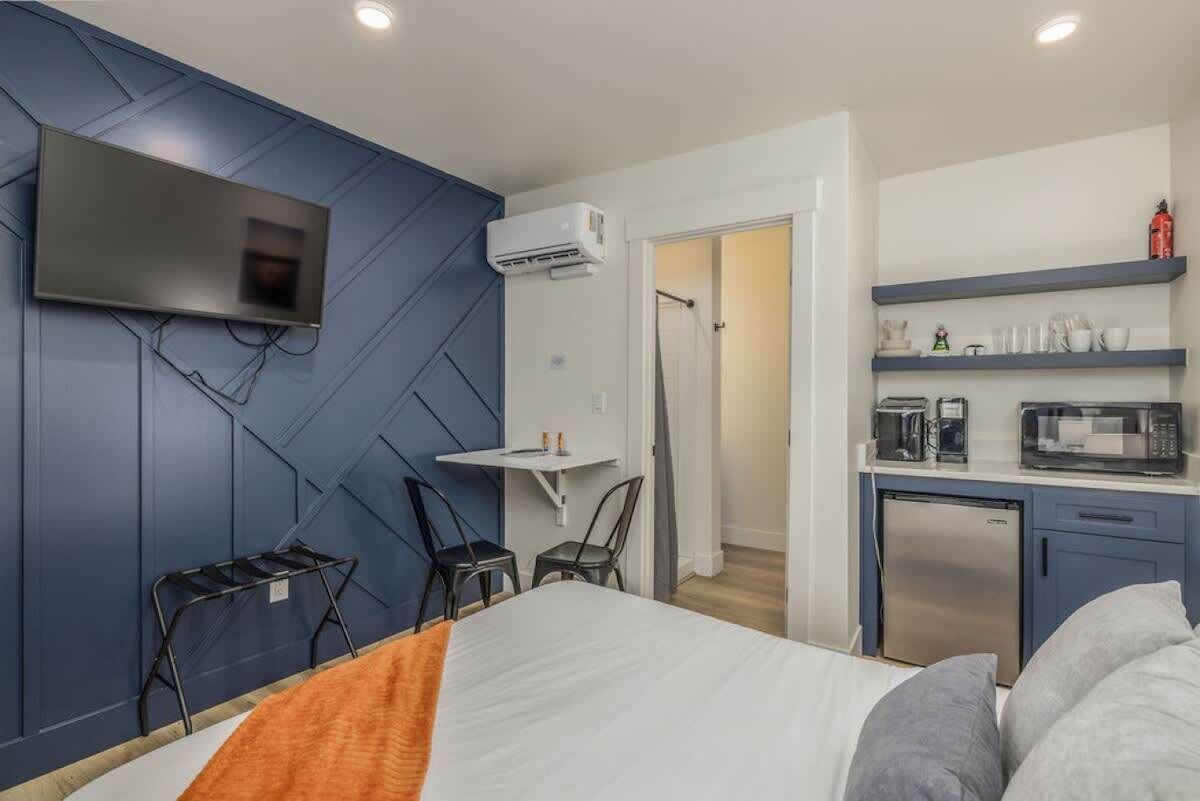 A modern hotel room with a bed, a TV mounted on the wall, a small table with chairs, a kitchenette with a microwave and coffee maker, and an air conditioning unit.