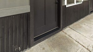Fachada oscura de un edificio con una puerta numerada, una ventana con marco blanco y un pequeño alero sobre la puerta.
