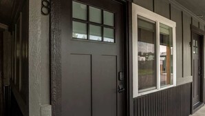 Une porte d’entrée de couleur sombre, surmontée d’une petite fenêtre, un numéro « 8 » bien visible sur le mur, et un chemin en béton menant à la porte.