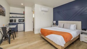 A modern bedroom with a wooden bed, an orange throw blanket, a white bedside table, and a nightstand. Adjacent is a compact kitchenette with a microwave, coffee maker, and small refrigerator. - Studio #3 At The Flats - Downtown Nightlife (Coeur d'Alene)