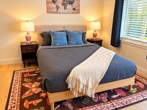 A bed with a dark blue mattress and a white throw blanket, flanked by two nightstands with lamps, a world map on the wall, and a window with blinds.