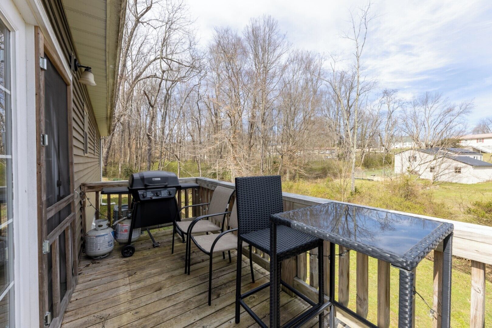 Eine Holzterrasse mit Grill, zwei Stühlen und einem Tisch mit Blick auf ein bewaldetes Gebiet.
