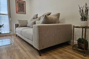A neutral-toned sofa with multiple cushions, a clear glass coffee table, and a potted plant on a side table.