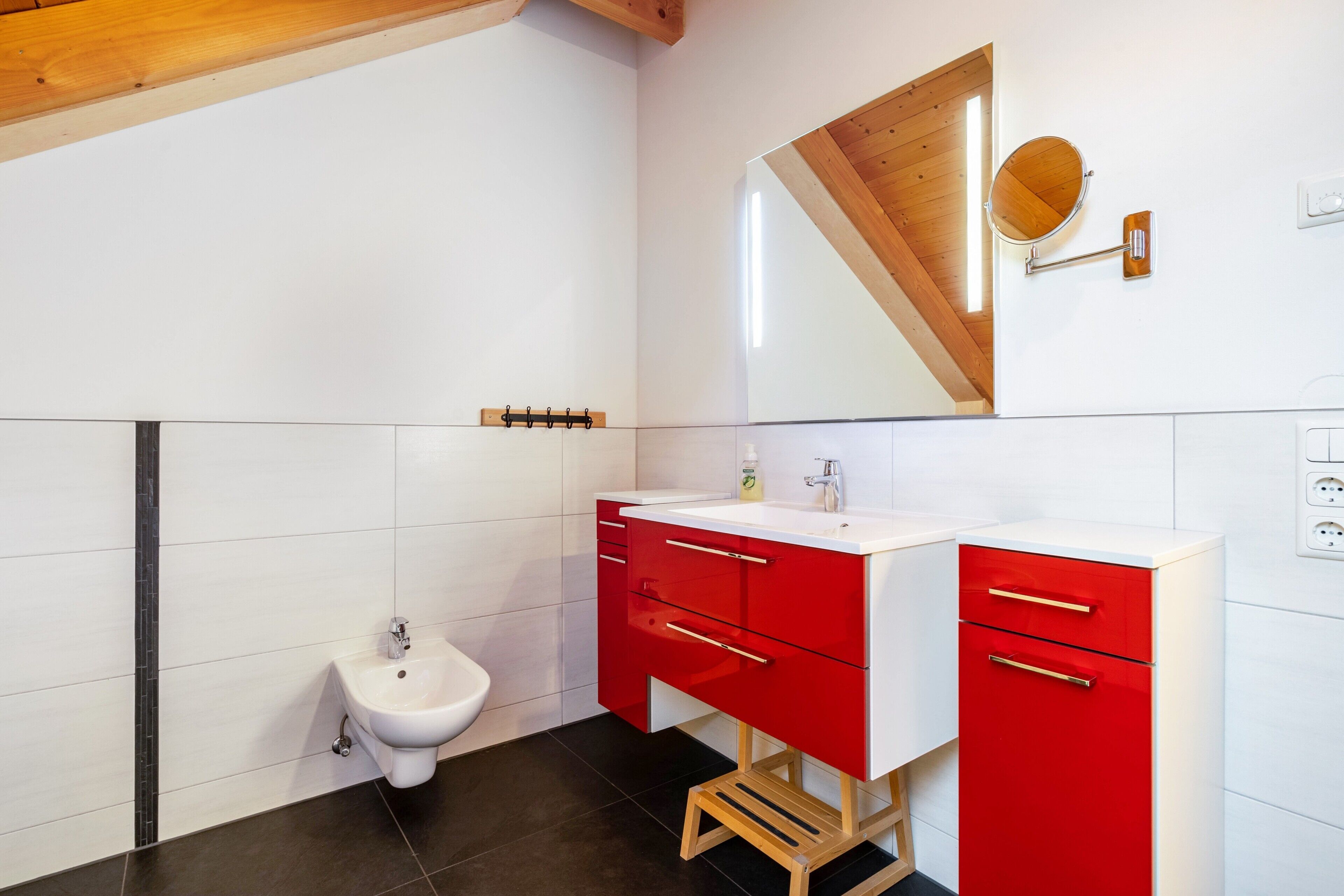 A modern bathroom with a bidet, a red vanity with drawers, a mirror, and a wooden ceiling beam.