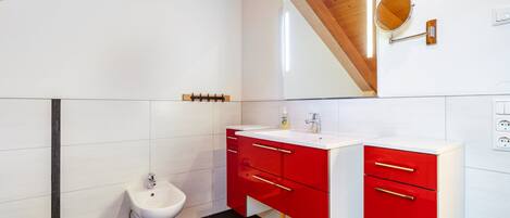 A modern bathroom with a bidet, a red vanity with drawers, a mirror, and a wooden ceiling beam.