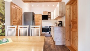 A kitchen with wooden cabinets, a refrigerator, an oven, and a dining area with chairs and a table. - Apartment 'Blockhaus - Ferienwohnung Sunset' with Shared Garden (Augustusburg-Erdmannsdorf)