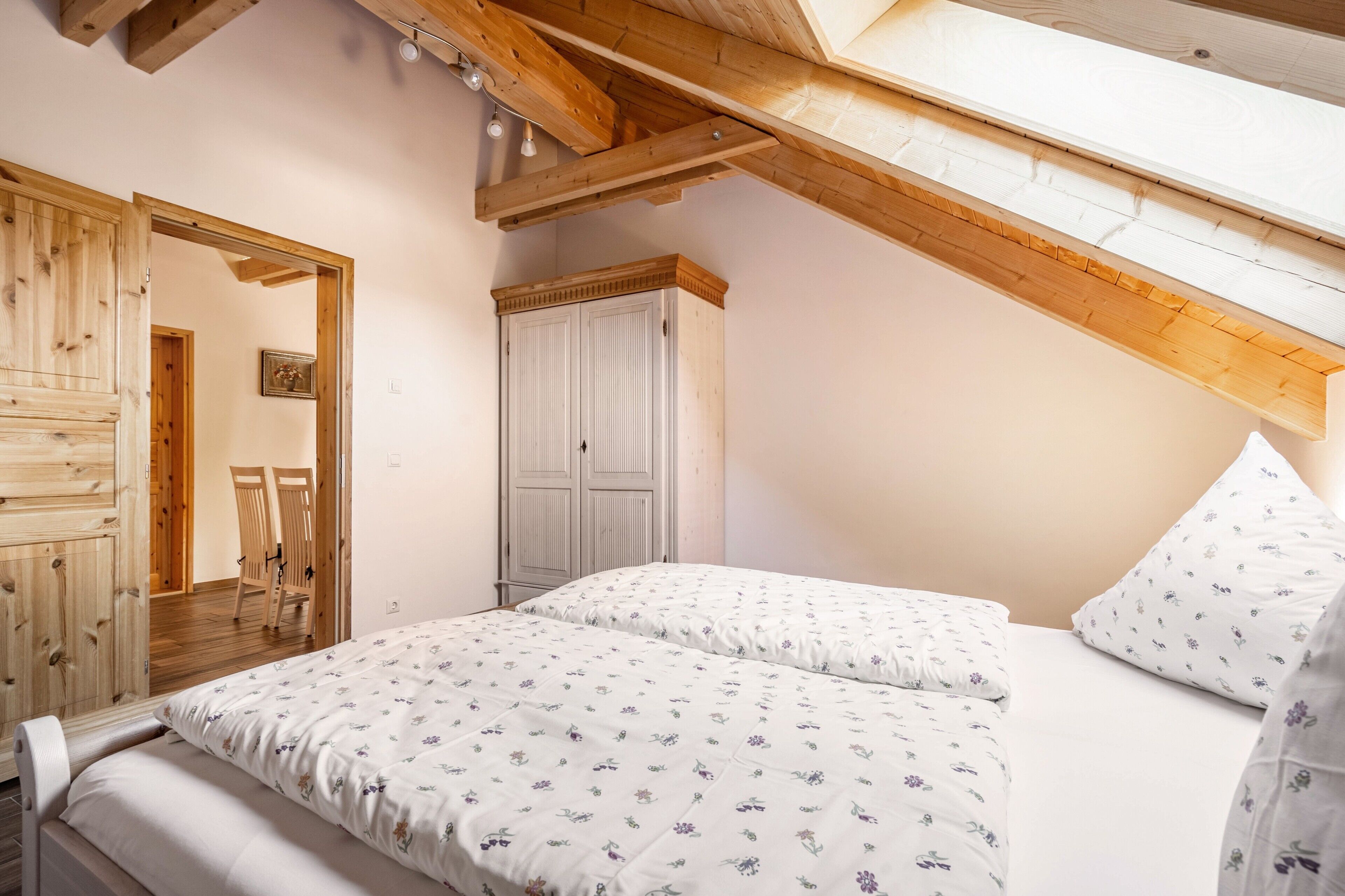 A bedroom with a sloped ceiling, wooden beams, a wardrobe, and a bed with floral bedding.