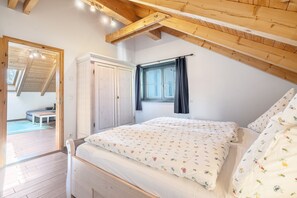 Ein Schlafzimmer mit schräg angesetzter Decke, einem Bett mit floralem Bettzeug, einem Kleiderschrank und einem Fenster mit Vorhängen.
