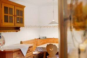 Une pièce comprenant un meuble en bois, une table recouverte d’une nappe à motifs floraux, une chaise en bois et un luminaire suspendu.