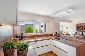 A modern kitchen with a large island, wooden countertops, white cabinetry, and a view of greenery outside.