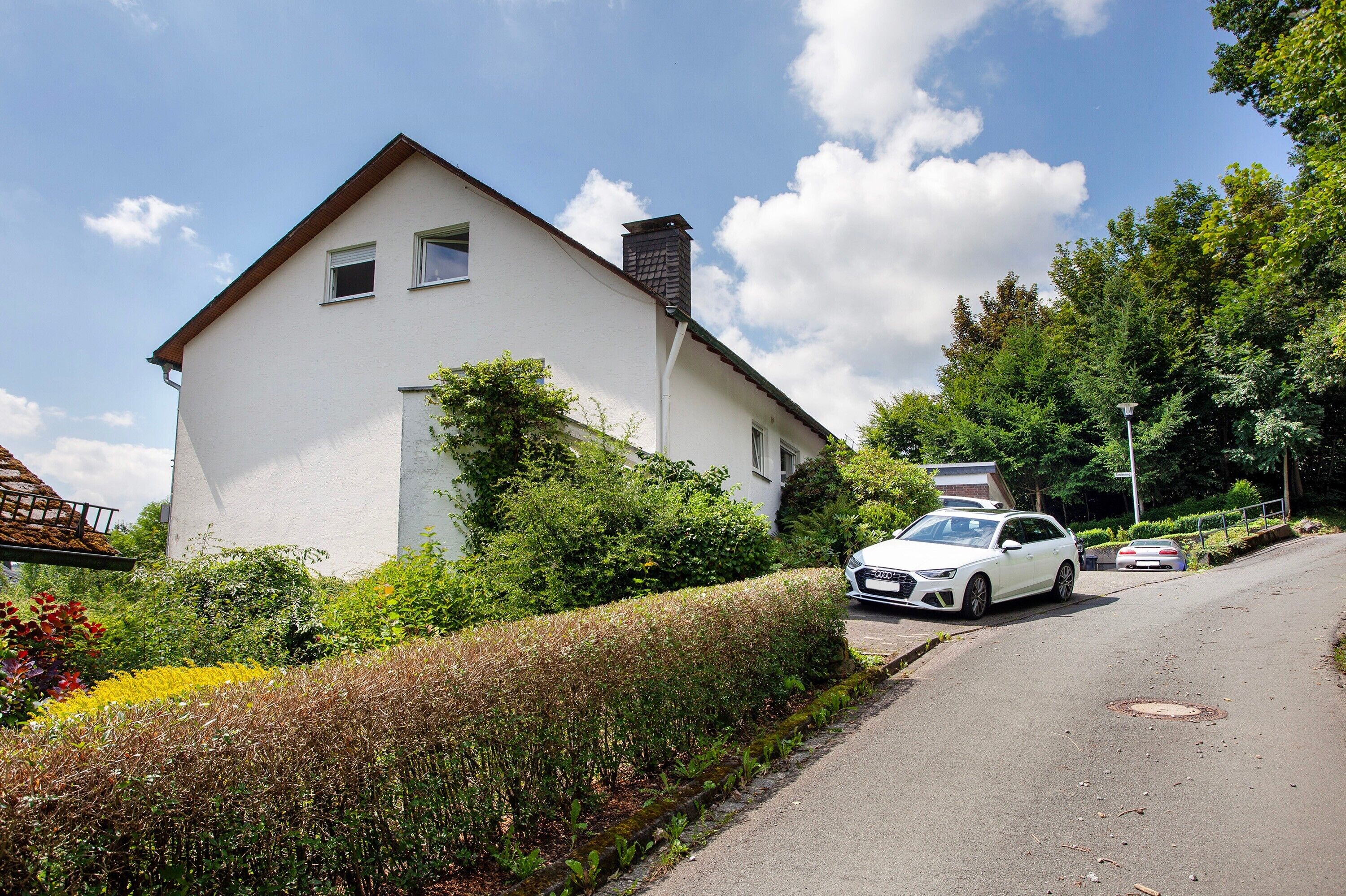 A white two-story house with a sloped roof, a chimney, and a car parked on the street.